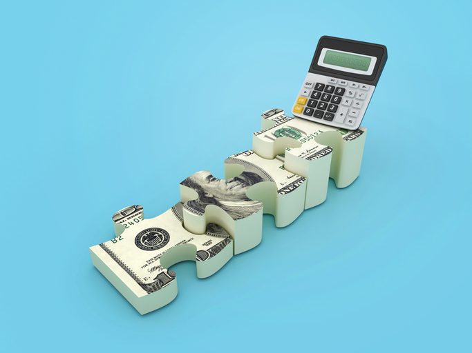A calculator sits on top of interlocking puzzle pieces designed to look like hundred‑dollar bills against a light blue background.