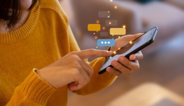 Photo of a woman with medium‑length dark hair wearing a mustard yellow sweater, using a smartphone indoors. She taps the screen with one hand while holding the phone in the other. Colorful illustrated chat bubbles float above the phone, symbolizing messaging or social interaction.