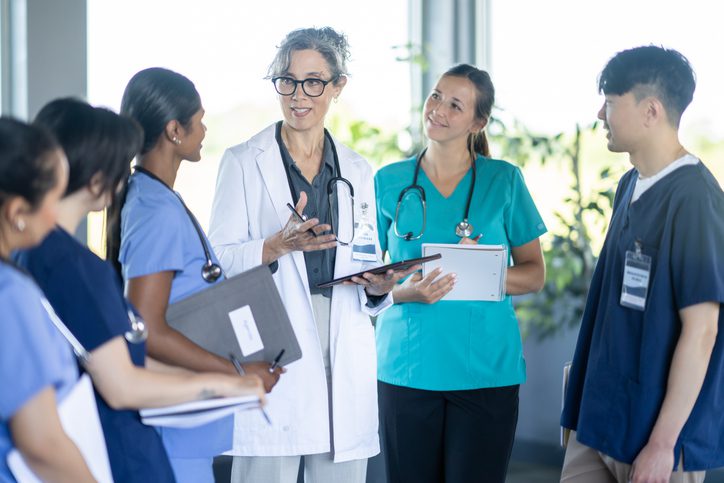 A small group of medical professionals gathers in a hospital hallway for a training discussion, listening closely as the lead physician.