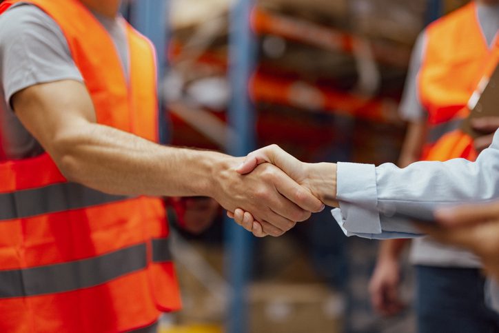 Photo of two people shaking hands in a warehouse. One man wears an orange safety vest and gray shirt; the other person wears a light blue long‑sleeve shirt. Shelving and another person in an orange vest are visible in the background.