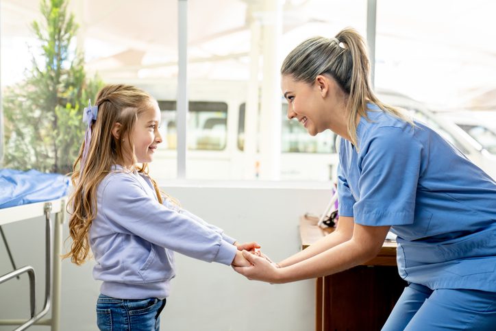 A healthcare worker in blue scrubs kneels to hold hands with a young girl student in a casual outfit, offering support in a bright school setting.