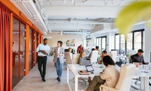 Photo of a modern open-plan office with several people working at desks and two individuals walking and talking in the foreground. The space features large windows, white walls, orange accents, and contemporary furniture.