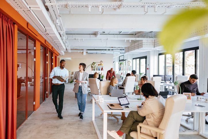 Photo of a modern open-plan office with several people working at desks and two individuals walking and talking in the foreground. The space features large windows, white walls, orange accents, and contemporary furniture.