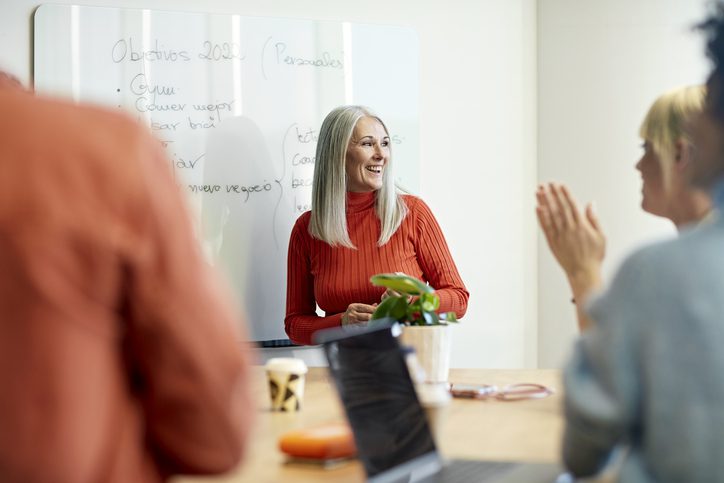 Photo of a woman with long gray hair wearing a red turtleneck sweater standing in front of a whiteboard, speaking to two people seated at a table. One person is clapping and another is partially visible in the foreground. Laptops, a mug, and a small plant are on the table.