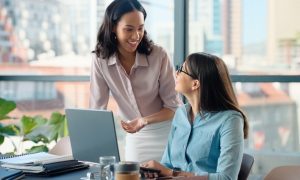 Two women collaborate at a modern office desk, with one standing and gesturing toward an open laptop while the other sits, listens, and learns. The workspace includes notebooks, a coffee cup, and a smartphone, and large windows behind them reveal a city skyline filled with natural light.