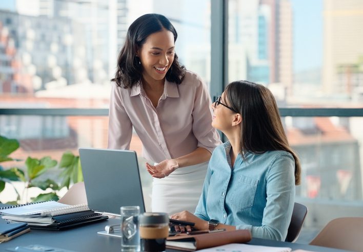 Two women collaborate at a modern office desk, with one standing and gesturing toward an open laptop while the other sits, listens, and learns. The workspace includes notebooks, a coffee cup, and a smartphone, and large windows behind them reveal a city skyline filled with natural light.