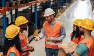 Photo of a group of workers in a warehouse wearing orange safety vests and yellow or white hard hats. A man in a white hard hat and orange vest stands at the center holding a tablet and speaking to the team. The others, including women and men, stand around him listening; one person on the right holds a cardboard box. Rows of shelves and inventory line the background.