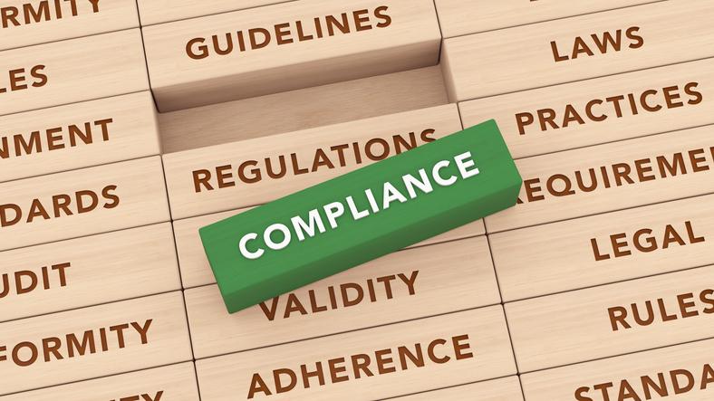Photo of a green rectangular block labeled “COMPLIANCE” placed diagonally on stacked wooden blocks engraved with words including “REGULATIONS,” “GUIDELINES,” “LAWS,” “REQUIREMENTS,” “LEGAL,” “RULES,” “STANDARDS,” “VALIDITY,” “ADHERENCE,” “CONFORMITY,” and “AUDIT.”
