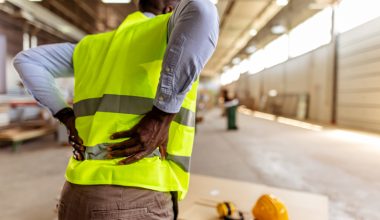 Photo of a person wearing a high-visibility yellow safety vest over a light blue long-sleeve shirt and brown pants, standing indoors in an industrial workspace with both hands placed on the lower back; a yellow hard hat and ear protection rest on a work surface in the foreground.