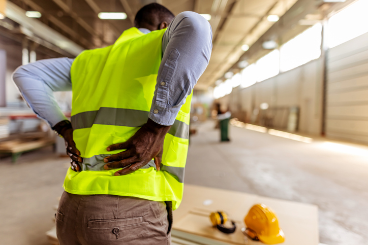 Photo of a person wearing a high-visibility yellow safety vest over a light blue long-sleeve shirt and brown pants, standing indoors in an industrial workspace with both hands placed on the lower back; a yellow hard hat and ear protection rest on a work surface in the foreground.