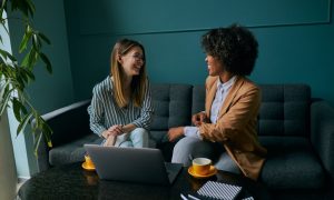 Two young businesswomen are networking in a quiet coffee shop, collaborating over a laptop in a calm, introvert‑friendly setting that supports building referrals without small talk.