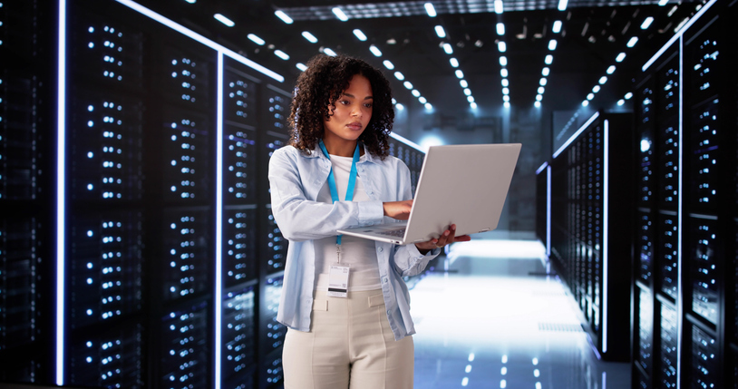 Photo: adult woman with curly dark hair wearing a light blue shirt, ID lanyard, and beige pants, using a laptop while standing in a data center aisle lined with illuminated server racks.