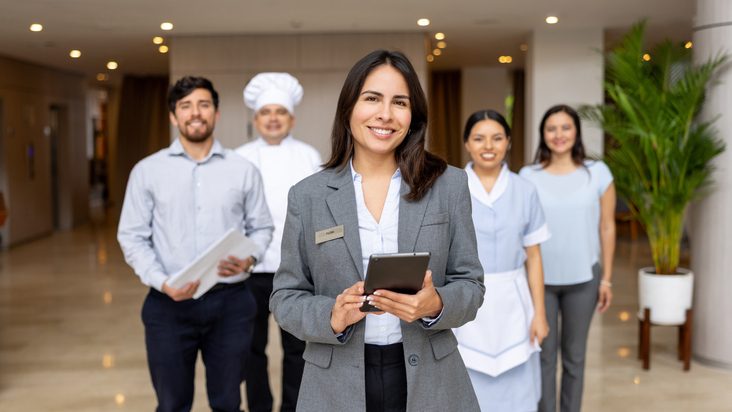 Photo of five hospitality staff standing in a hotel lobby; a person in a gray blazer with a name badge holds a tablet in the foreground, with colleagues behind wearing business attire, a chef’s uniform, and a housekeeping uniform. Large potted plant and softly lit interior in the background.