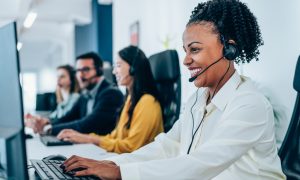 Photo of customer service representative wearing a white button-down shirt and headset, typing at a computer workstation in a modern call center, with three additional headset-wearing colleagues and monitors visible in the background.