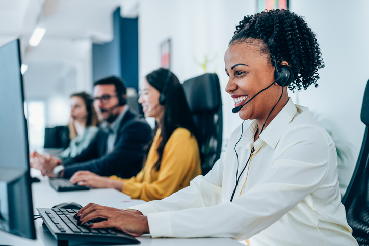 Photo of customer service representative wearing a white button-down shirt and headset, typing at a computer workstation in a modern call center, with three additional headset-wearing colleagues and monitors visible in the background.