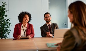 Photo of a professional interview or meeting in a conference room, with two people seated behind a wooden table, one holding a tablet and another seated beside an open laptop, facing a third person in the foreground. Glass door, potted plant, and modern office setting visible in the background.