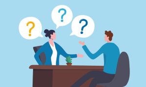 Illustration of two office workers seated across a desk in conversation, with speech bubbles containing question marks above them, symbolizing a discussion, interview, or Q&A session; blue background, brown desk, and small potted plant on the tabletop.