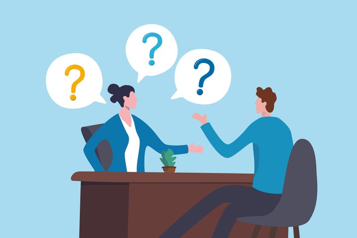 Illustration of two office workers seated across a desk in conversation, with speech bubbles containing question marks above them, symbolizing a discussion, interview, or Q&A session; blue background, brown desk, and small potted plant on the tabletop.