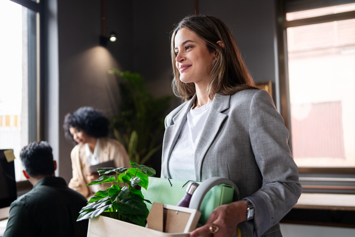 Photo of a person in a light gray blazer and white shirt carrying a box with a green plant and office supplies in a modern office; two other people are blurred in the background near desks and windows.